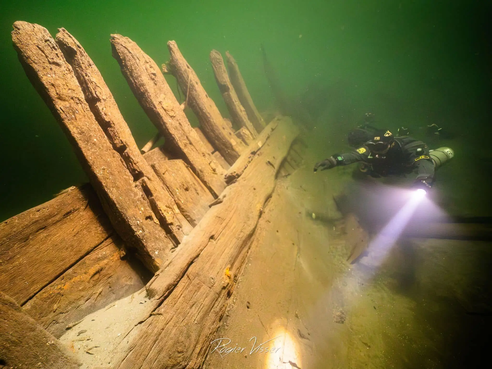 Houten scheepswrak met opstaande spanten in groen binnenwater, een duiker met lamp ernaast